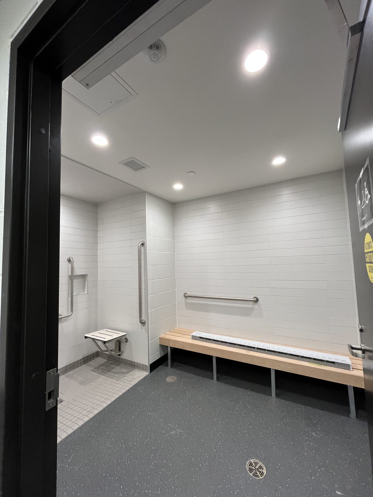 Accessible shower and change room with white tile walls, grab bars, a fold-down seat and a wood bench at the Albert McCormick Community Centre in Waterloo