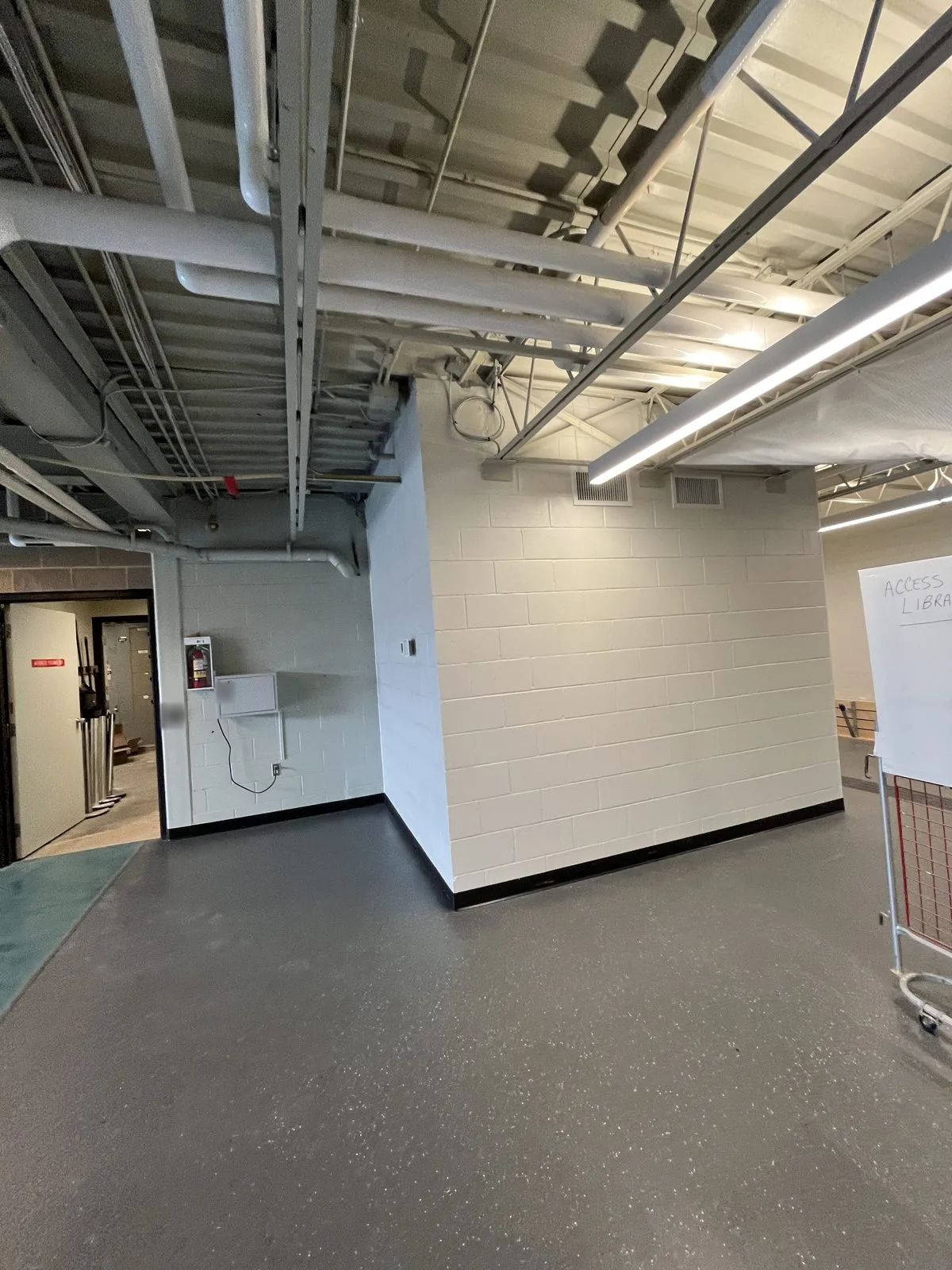 Lower-level corridor with white painted block walls, black base and white painted exposed ceiling and piping at the Albert McCormick Community Centre in Waterloo