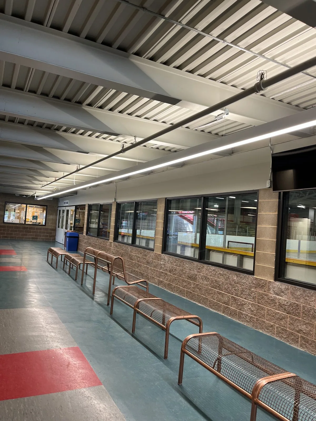 Rink viewing corridor with white painted steel deck ceiling, split-face block walls, rink viewing windows and bronze mesh benches at the Albert McCormick Community Centre in Waterloo