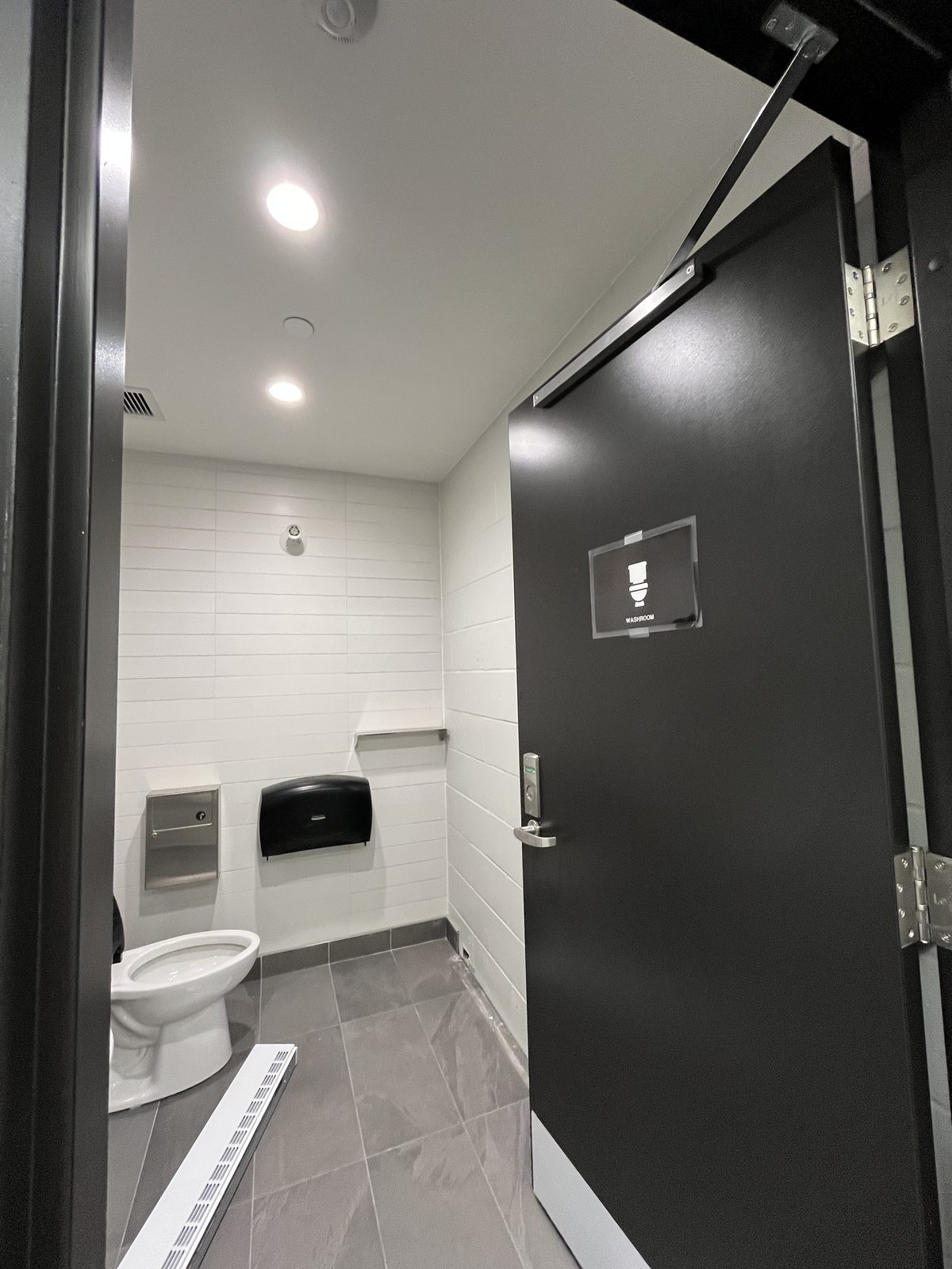 Single-user washroom with white tile walls, grey floor tile and a black door at the Albert McCormick Community Centre in Waterloo