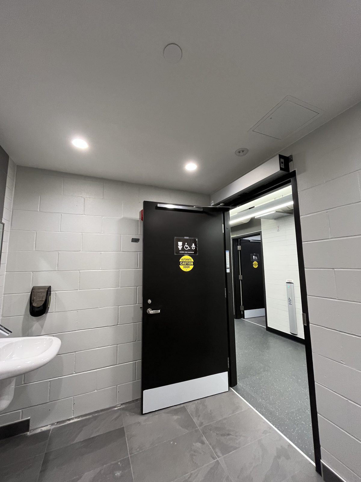 Barrier-free washroom entrance with a black automatic door, white painted block walls and grey tile floor at the Albert McCormick Community Centre in Waterloo