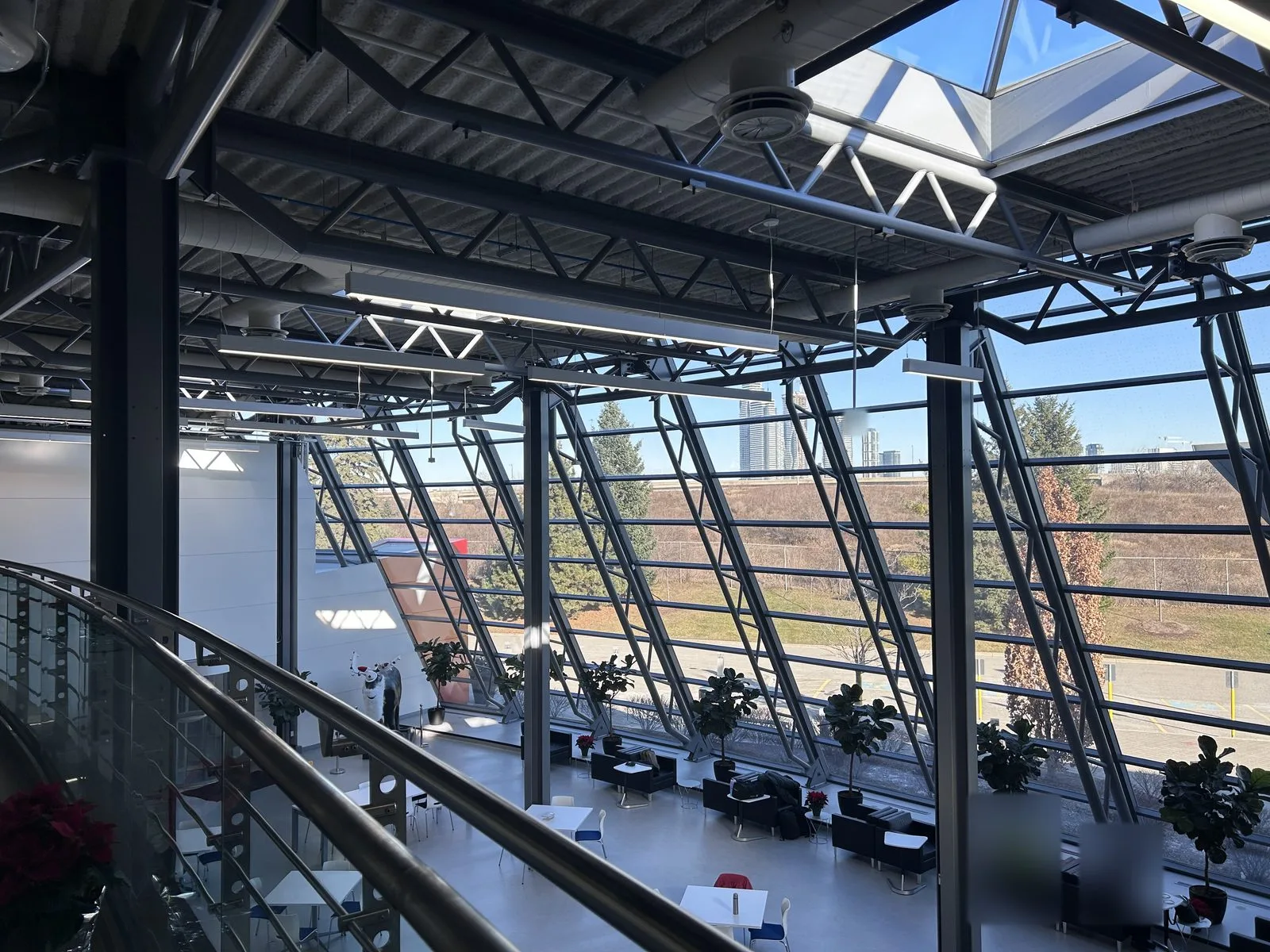 View from the mezzanine over a sunlit atrium lounge with a sloped glazed curtain wall and exposed painted steel trusses