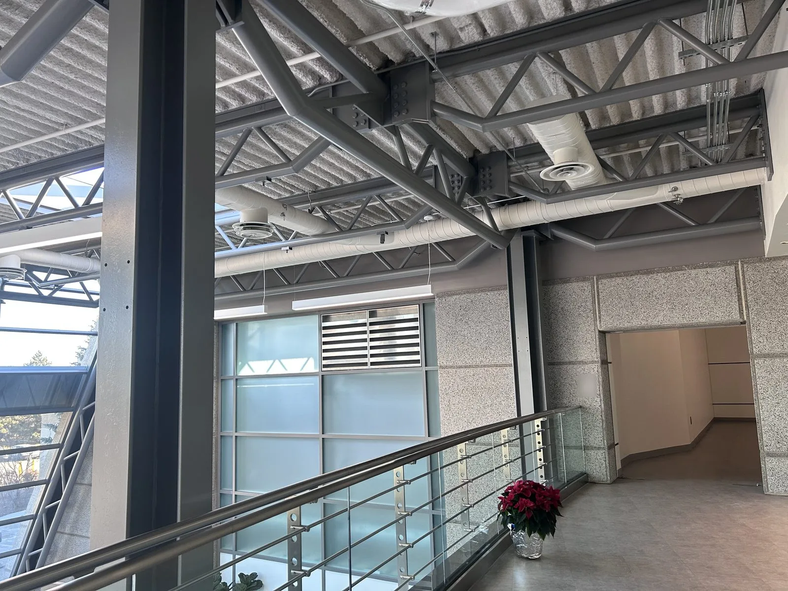 Mezzanine walkway with glass railing, grey painted steel column and exposed trusses leading to a corridor opening