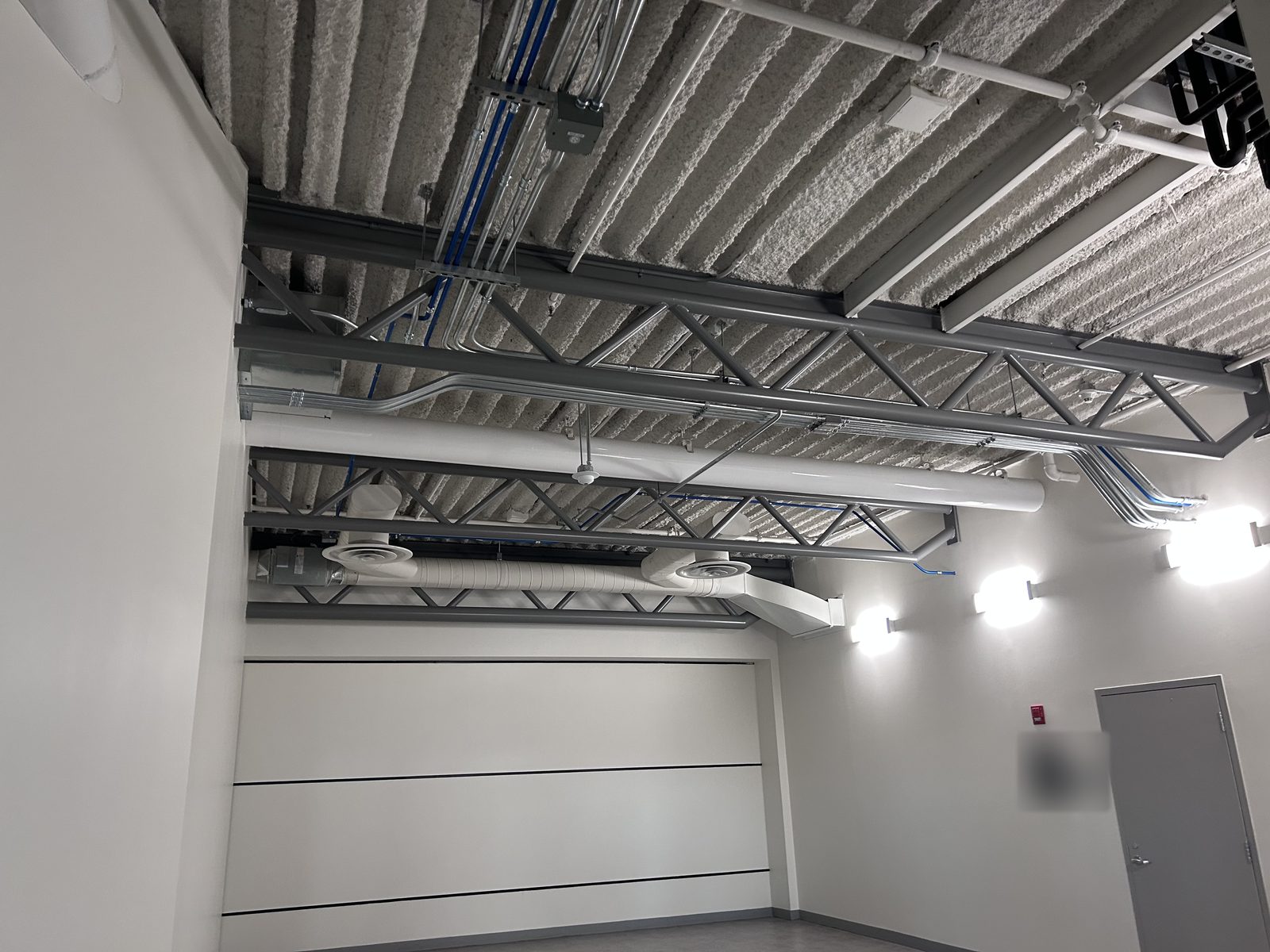 Room with white painted walls, exposed painted steel joists, ductwork and wall sconces beside a grey door