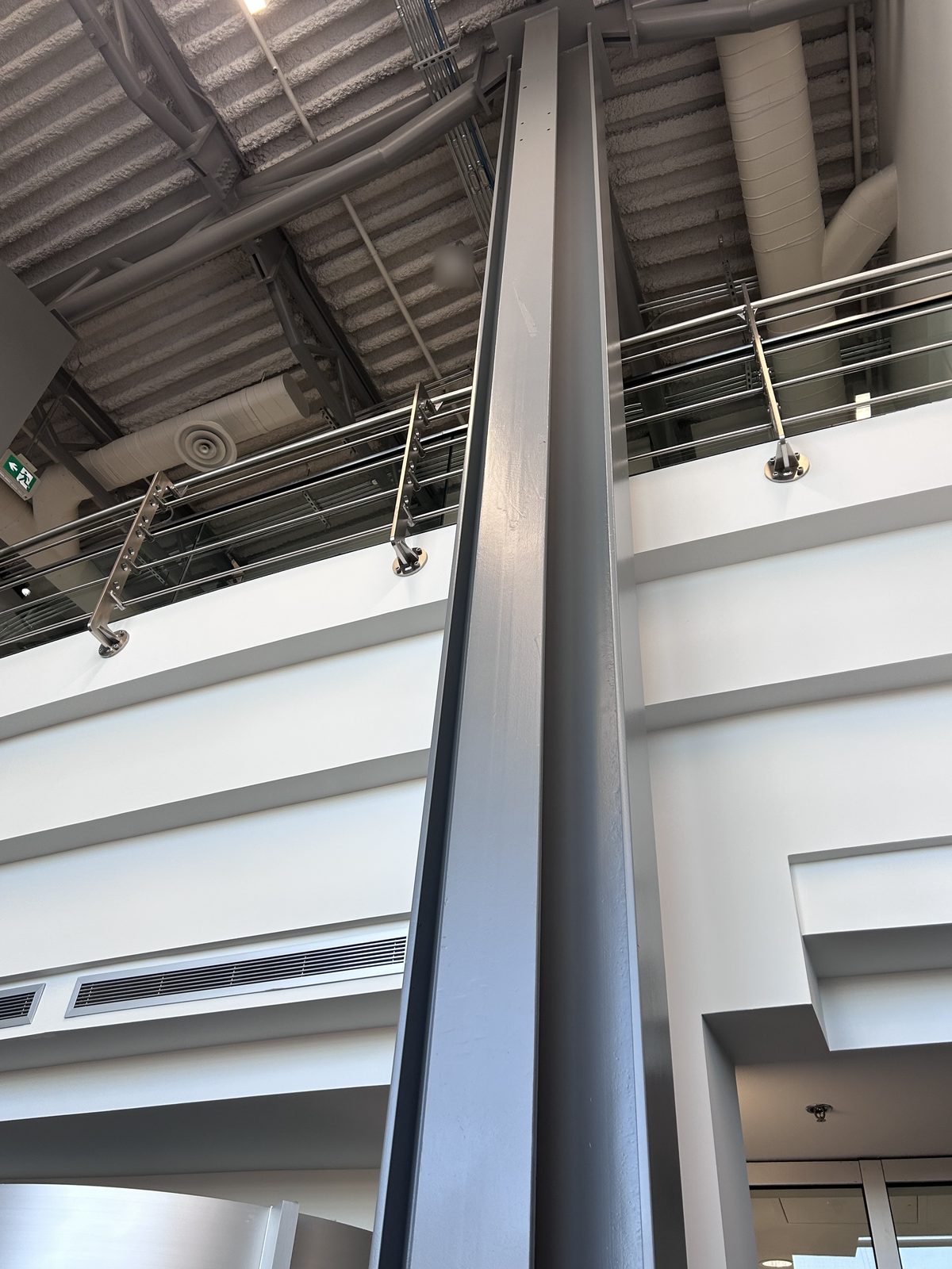 Grey painted steel column rising past white mezzanine fascias with stainless glass railings