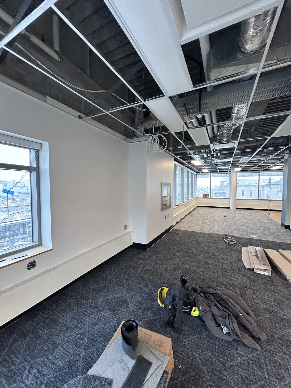 White painted walls and a window in a carpeted room with an open ceiling grid and tools on the floor