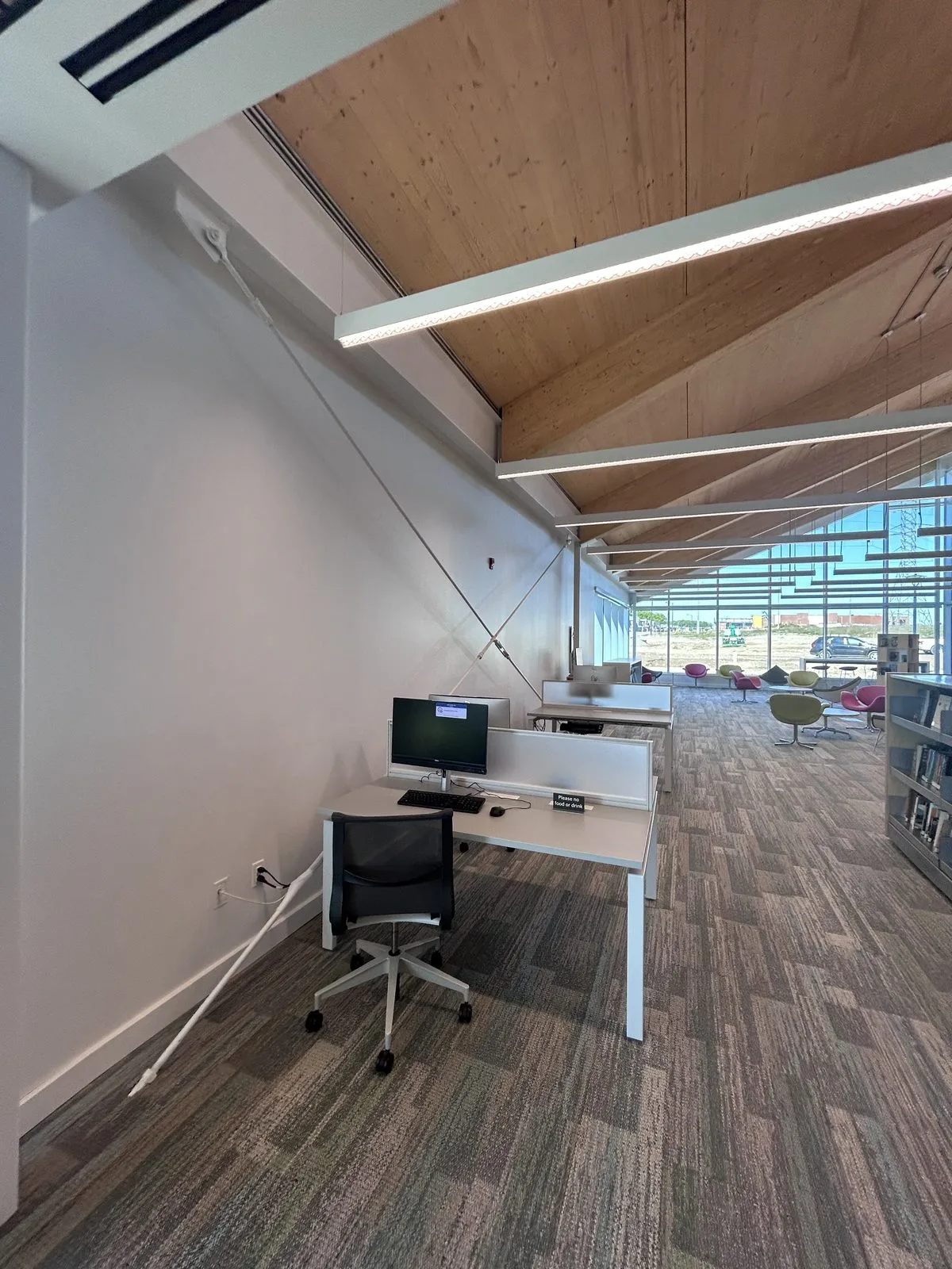 Computer stations along a white wall beside a lounge with colourful chairs and a glazed end wall