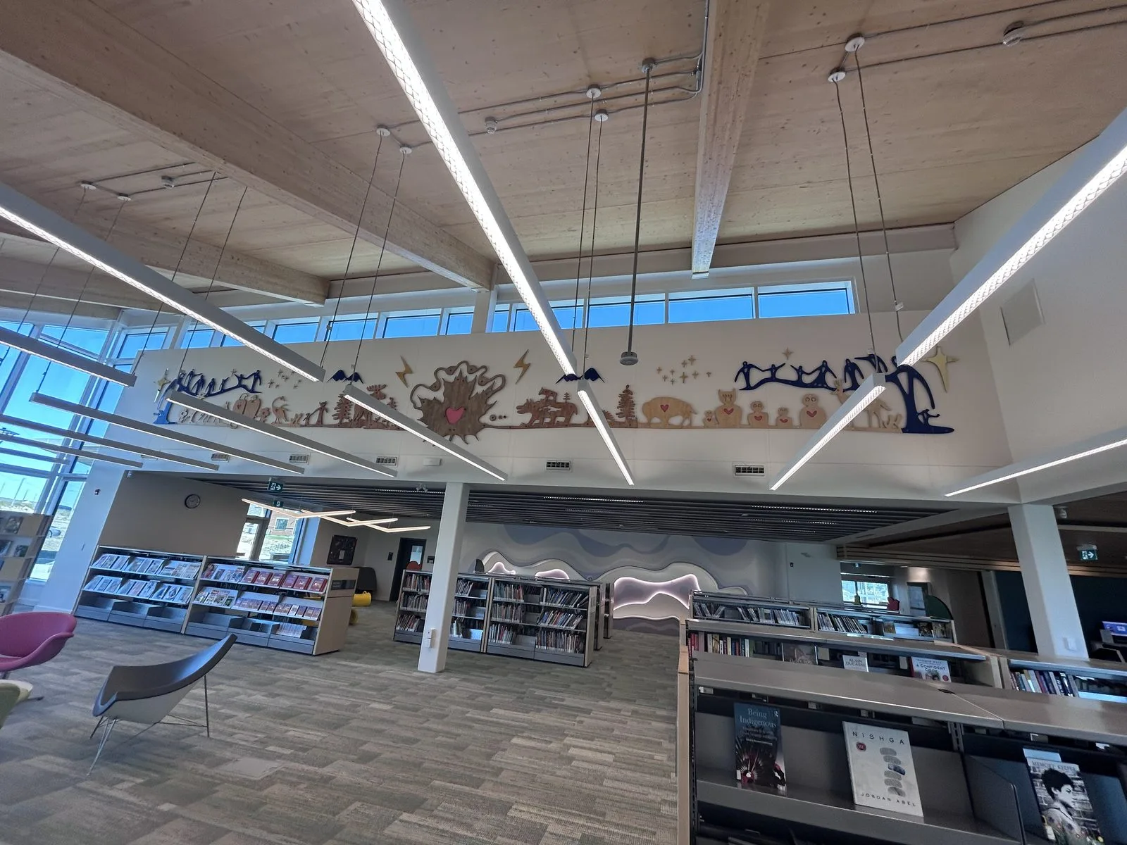 Main library hall with a large cut-out mural frieze, exposed timber ceiling, linear pendant lights and book stacks