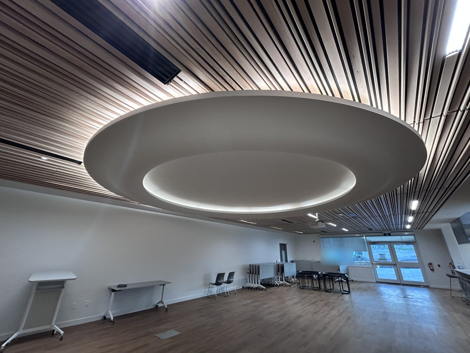 Multipurpose room with an oval ceiling feature in a wood slat ceiling, folded tables and double exit doors